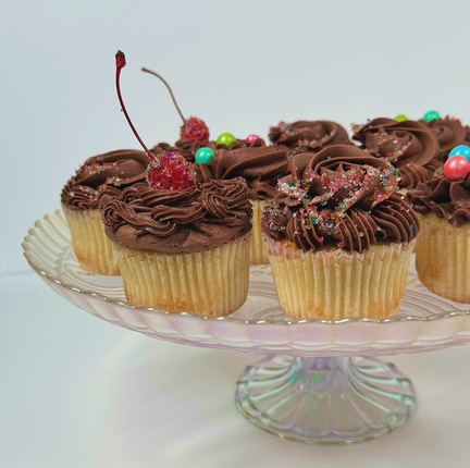 Several yellow-cake cupcakes with rich chocolate buttercream on a glass cake stand.  The cupcakes feature various sprinkles, finishing sugars, and glitter-dipped cherries as toppings.