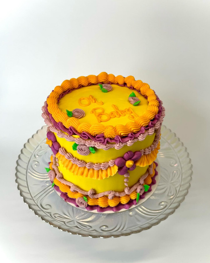 An 8 inch 3 layer vanilla cake with lemon curd filling. The cake is decorated in a whimsical vintage style with a yellow base color, purple, lavender and orange piping with floral frosting designs.  It says "Oh Baby" on top for a baby shower.